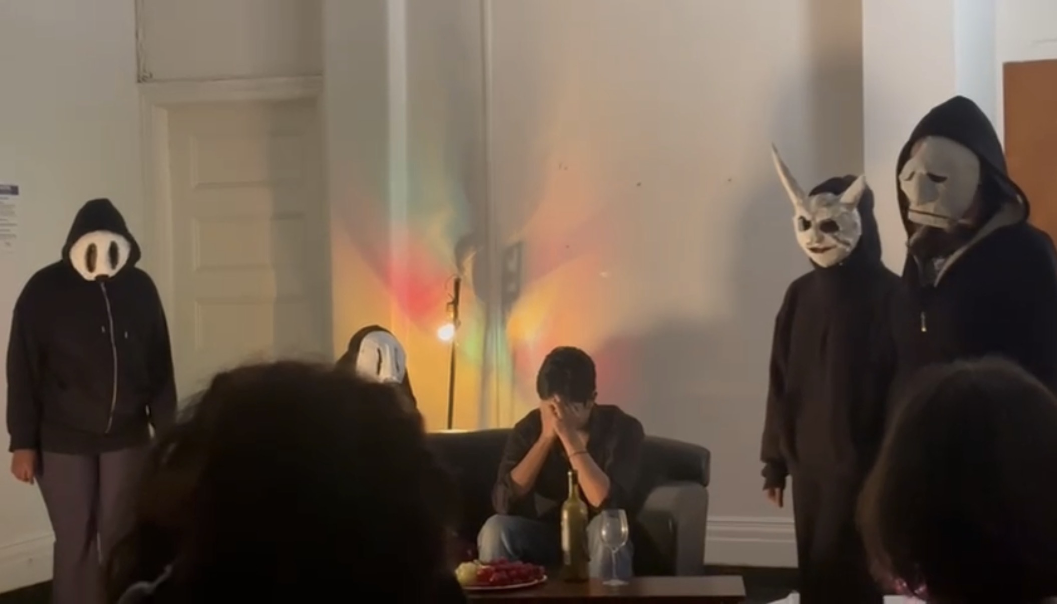 Masked student performers surrounding a seated performer during the first play that inspired the festival