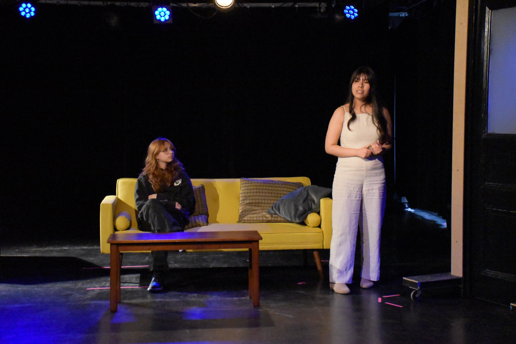Two student performers in a scene with a yellow couch on a black box stage
